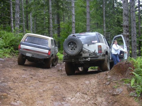 1991 Ford Bronco - Two off-road vehicles navigate a muddy path through a dense forest.