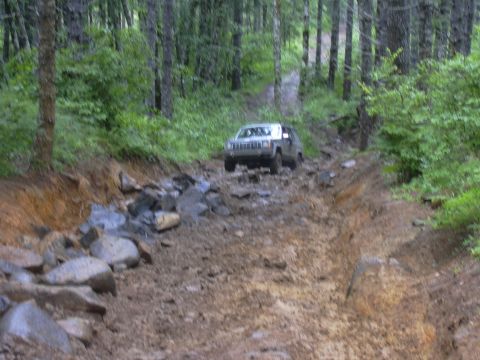 1991 Ford Bronco - A Jeep Cherokee XJ navigates a challenging muddy and rocky off-road trail through a dense forest.