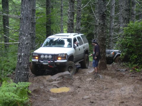 1991 Ford Bronco - Modified SUVs tackle a challenging muddy trail in a wooded area.