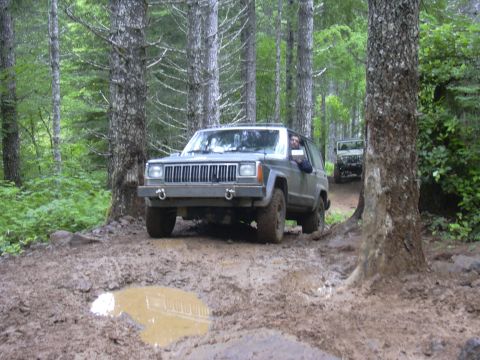 1991 Ford Bronco - Two Jeep Cherokees navigate a muddy path in a dense forest during an off-road excursion.