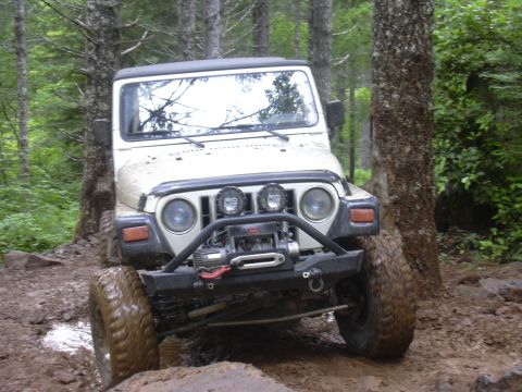 1991 Ford Bronco - A Jeep Wrangler navigates a challenging, muddy off-road path through a forest.