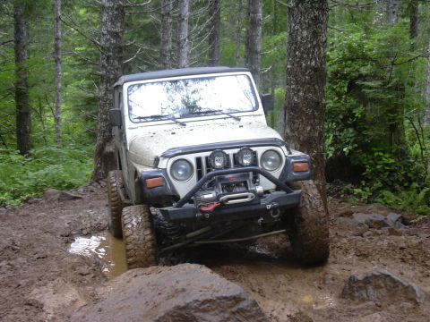 1991 Ford Bronco - A white Jeep Wrangler navigates a challenging muddy off-road trail through a forest.