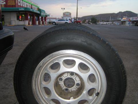1985 Ford Mustang - A close-up of a car's wheel and tire is shown with a country store and other vehicles in the background.