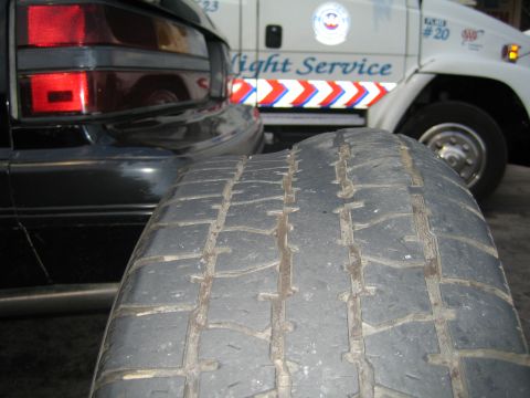 1985 Ford Mustang - A close-up of a worn tire is in the foreground, with the rear of a black car and a white service van visible behind it.