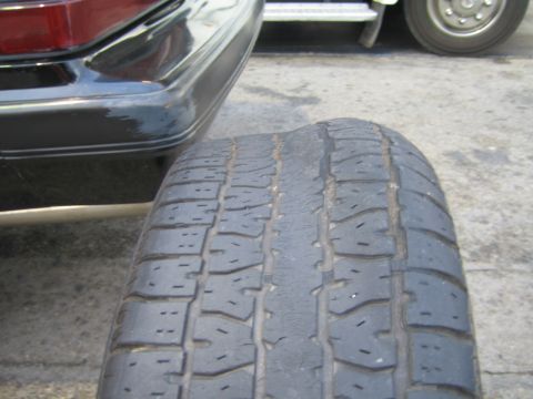 1985 Ford Mustang - A close-up shot focuses on a worn tire, with the rear bumper of a car visible to the left and another vehicle in the background.