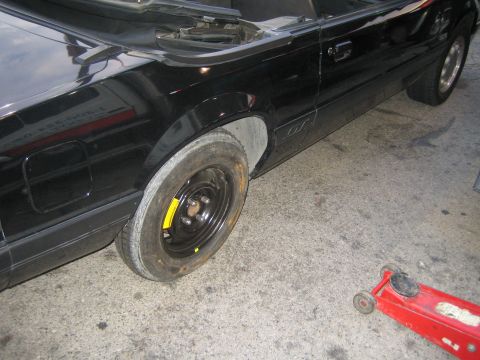 1985 Ford Mustang - A black 1985 Ford Mustang convertible is shown with a flat tire and a red jack on the ground nearby.