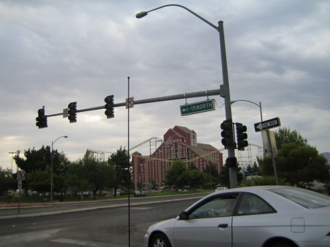 1985 Ford Mustang - A silver coupe drives through an intersection with a large hotel and roller coaster visible in the background under a cloudy sky.