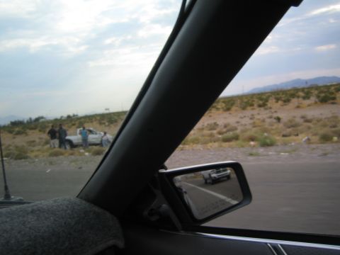 1985 Ford Mustang - A group of people are gathered around a white pickup truck on a desolate roadside, with a car visible in the side mirror.