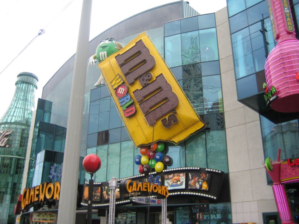 Giant yellow M&M's candy wrapper sign with M&M characters, a large Coca-Cola bottle sculpture, and a pink neon sign