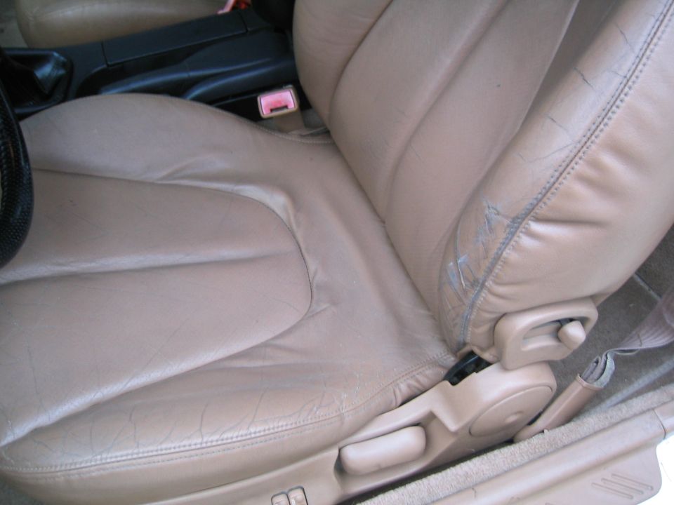 Tan leather car seat with visible wear and tear on the side bolster. Part of the center console and seat adjustment controls are visible