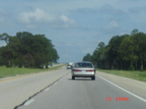 1988 Ford LTD Crown Victoria - A sedan travels down a highway under a cloudy sky, with trees lining the road.