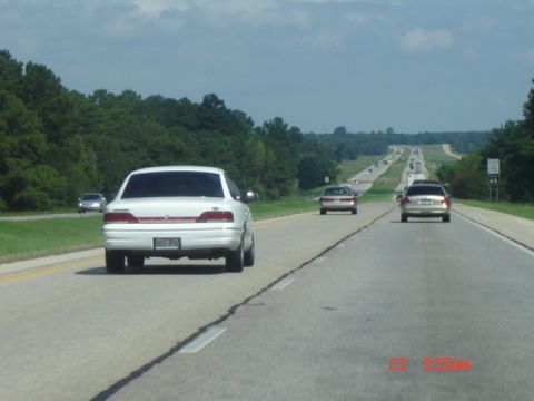 1988 Ford LTD Crown Victoria - A white sedan travels down a long, straight highway with trees lining the sides.