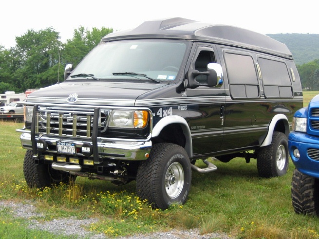 Black lifted Ford 4x4 conversion van with brush guard and off-road tires in a grassy field.