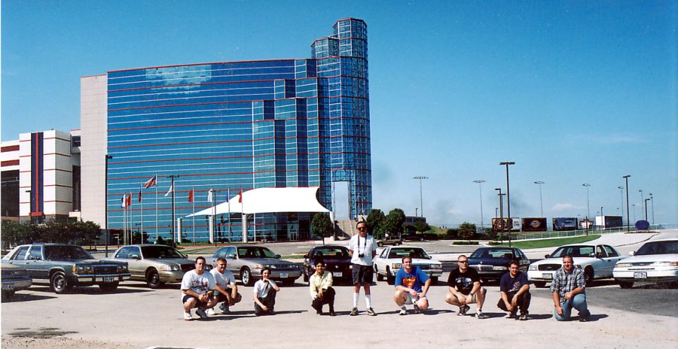 Group of people crouched in front of a row of full-size sedans by a blue glass high-rise