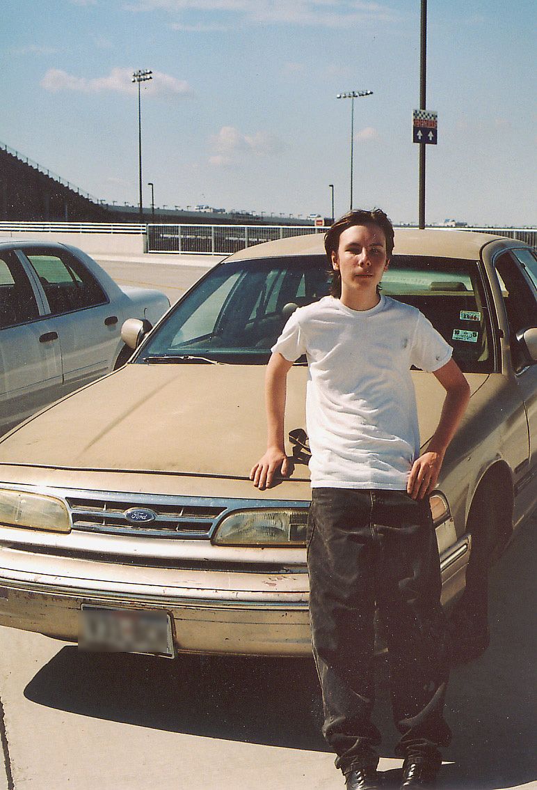 Teen in white shirt leaning on the front of a tan Ford Crown Victoria at a speedway lot