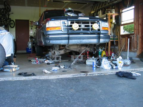 1995 Ford Bronco - Maroon and gray two-tone Ford Bronco up on jack stands in a home garage mid front-axle job, with the passenger front wheel off exposing the…