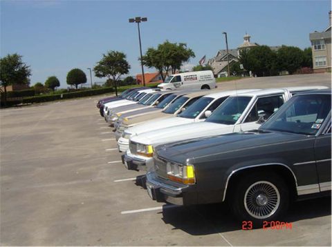 1988 Ford LTD Crown Victoria - Several sedans are parked in a lot under a clear blue sky, with buildings and trees in the background.
