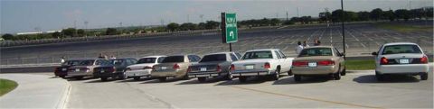 1988 Ford LTD Crown Victoria - A line of sedans, including a white 1988 Ford Crown Victoria LTD, are parked in a large lot near a race track.