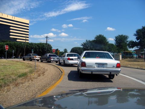 2000 Ford Crown Victoria - Several cars are driving down a road on a sunny day with a large building in the background.