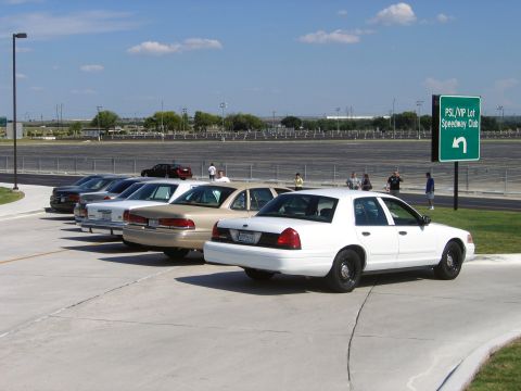 2000 Ford Crown Victoria - A line of sedans, likely police interceptors, are parked in a lot with a speedway in the background.