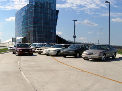 2000 Ford Crown Victoria - A line of sedans, including a maroon Ford Crown Victoria, are parked in a lot with a modern glass building in the background.