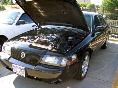 2000 Ford Crown Victoria - A black Mercury Grand Marquis is shown with its hood open, revealing the engine bay. Another vehicle is partially visible to the left.