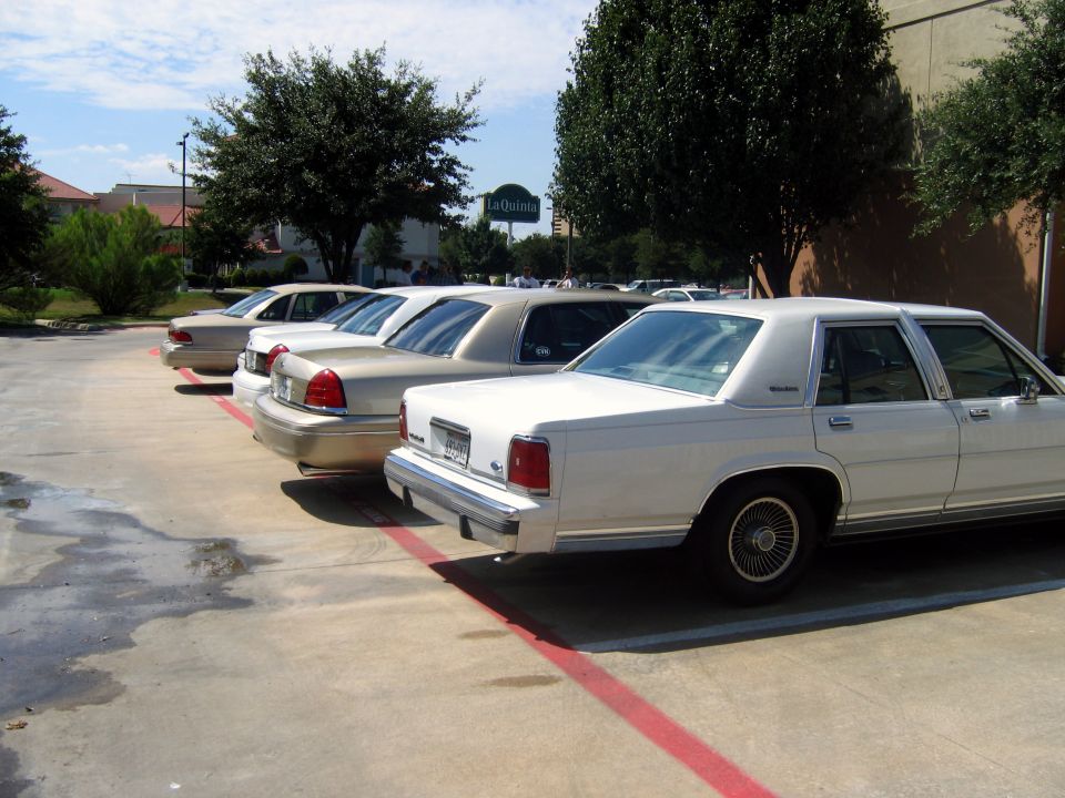 Four sedans parked in a lot, three are tan and one is white. The white one is closest to the camera