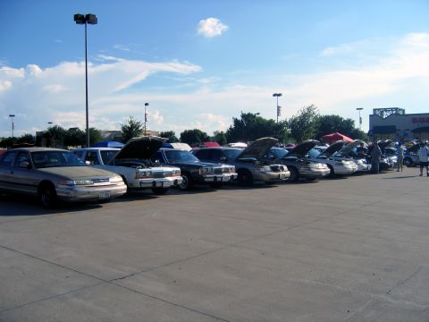 2000 Ford Crown Victoria - A gathering of Ford Crown Victorias, many with hoods open, parked in a lot under a blue sky.