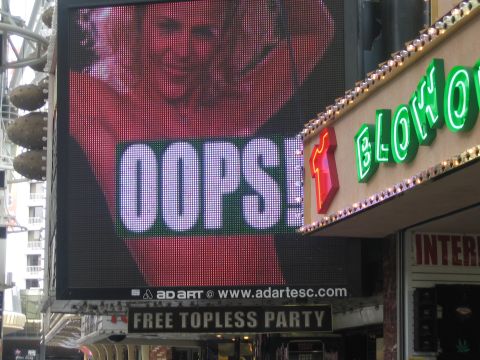 1985 Ford Mustang - A digital billboard displays 'OOPS!' next to a neon sign for 'BLOW' and a sign for a 'FREE TOPLESS PARTY'.