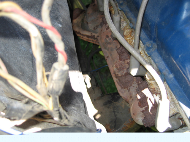 Engine bay close-up of a rusty exhaust manifold and aged wiring harness beside a blue fender