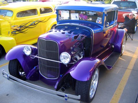 2000 Ford Crown Victoria - A vibrant purple hot rod with custom flame paintwork is parked next to a yellow classic car.