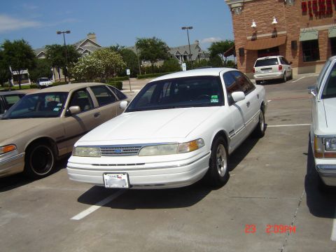 1988 Ford LTD Crown Victoria - A white sedan is parked in a lot next to another car, with a building in the background.