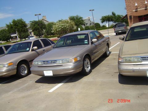 1988 Ford LTD Crown Victoria - Multiple tan sedans are parked in a lot under a bright sky.