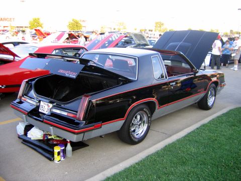 2000 Ford Crown Victoria - A black and gray coupe with red trim is displayed with its trunk and hood open at a car show, with other vehicles visible.