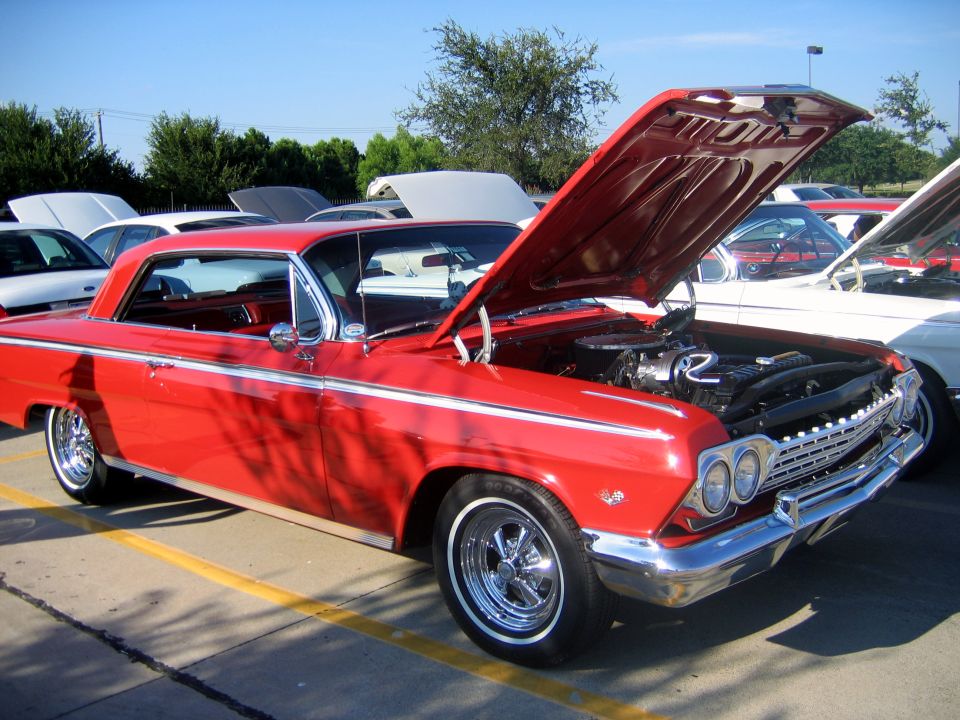 Red 1962 Chevrolet Impala coupe parked at a car show, hood open revealing engine. Other cars visible in background
