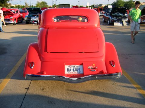2000 Ford Crown Victoria - A bright red coupe hot rod is parked at an outdoor car show, with other vehicles visible in the background.