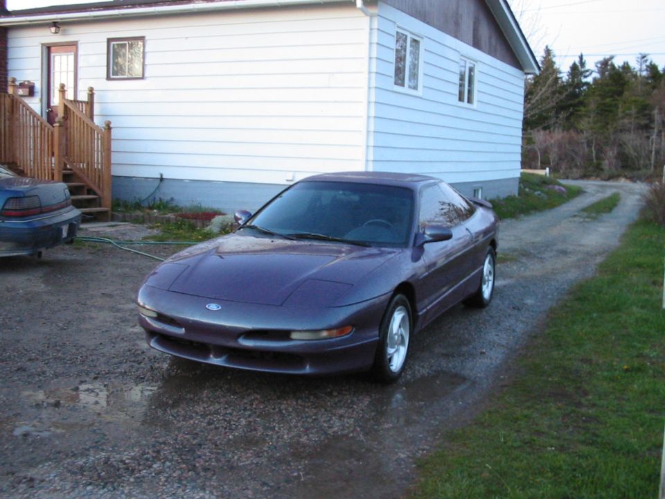 Dark purple Ford Probe coupe parked on wet gravel driveway beside a white house