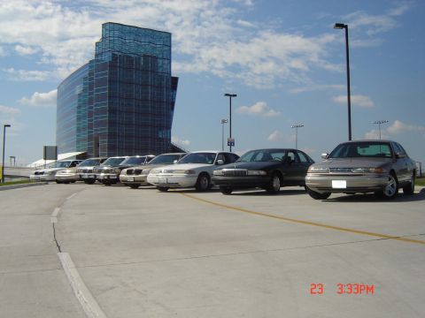 1988 Ford LTD Crown Victoria - A row of 1988 Ford Crown Victoria LTD sedans are parked in front of a distinctive glass building.