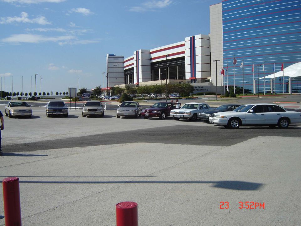 Several sedans parked in a lot with a large building in the background. The white sedan on the right appears to be a 1998 Ford Crown Victoria