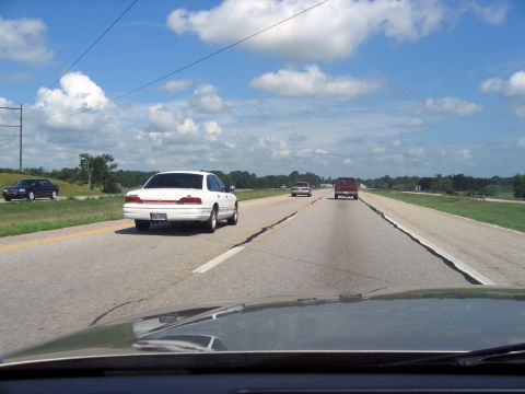 2000 Ford Crown Victoria - A white sedan drives down a highway on a sunny day with a few clouds in the sky.