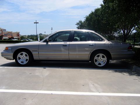 2000 Ford Crown Victoria - Driver-side profile of a light gold/champagne Ford Crown Victoria full-size sedan parked in a striped lot outside a strip mall, with amber…