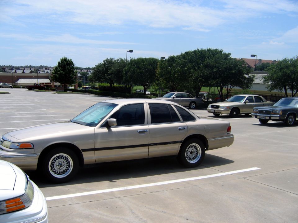 Tan sedan parked in a lot, side view. Multiple cars are visible in the background