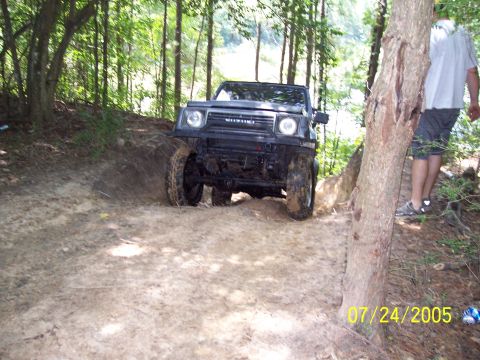 1987 Ford Bronco - Suzuki Samurai navigates a dirt path through a wooded area on a summer day.