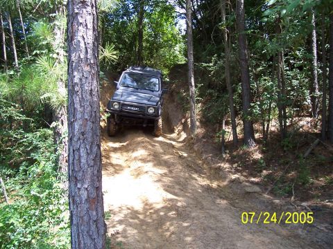 1987 Ford Bronco - A Suzuki Samurai navigates a steep, muddy trail surrounded by trees.