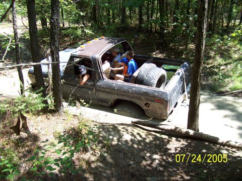 1987 Ford Bronco - A group of people are stuck in a muddy off-road vehicle in a wooded area.