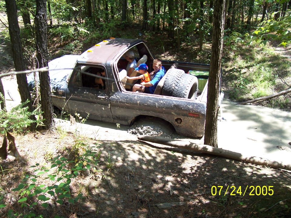 Brown 1978 Ford Bronco stuck in mud, people inside, spare tire in bed