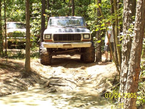 1987 Ford Bronco - A lifted Ford Bronco navigates a muddy path in a wooded area, with another vehicle visible in the background.