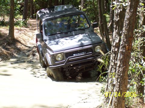 1987 Ford Bronco - Suzuki Samurai navigates a challenging muddy off-road course surrounded by trees.