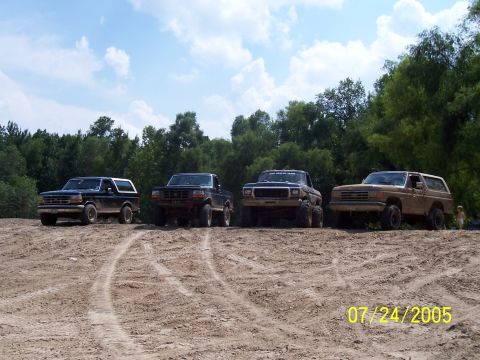 1987 Ford Bronco - A group of four-wheel drive vehicles are lined up on a dirt track under a partly cloudy sky.
