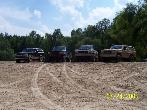 1987 Ford Bronco - Four 1987 Ford Bronco SUVs are lined up on a dirt track on a sunny day.